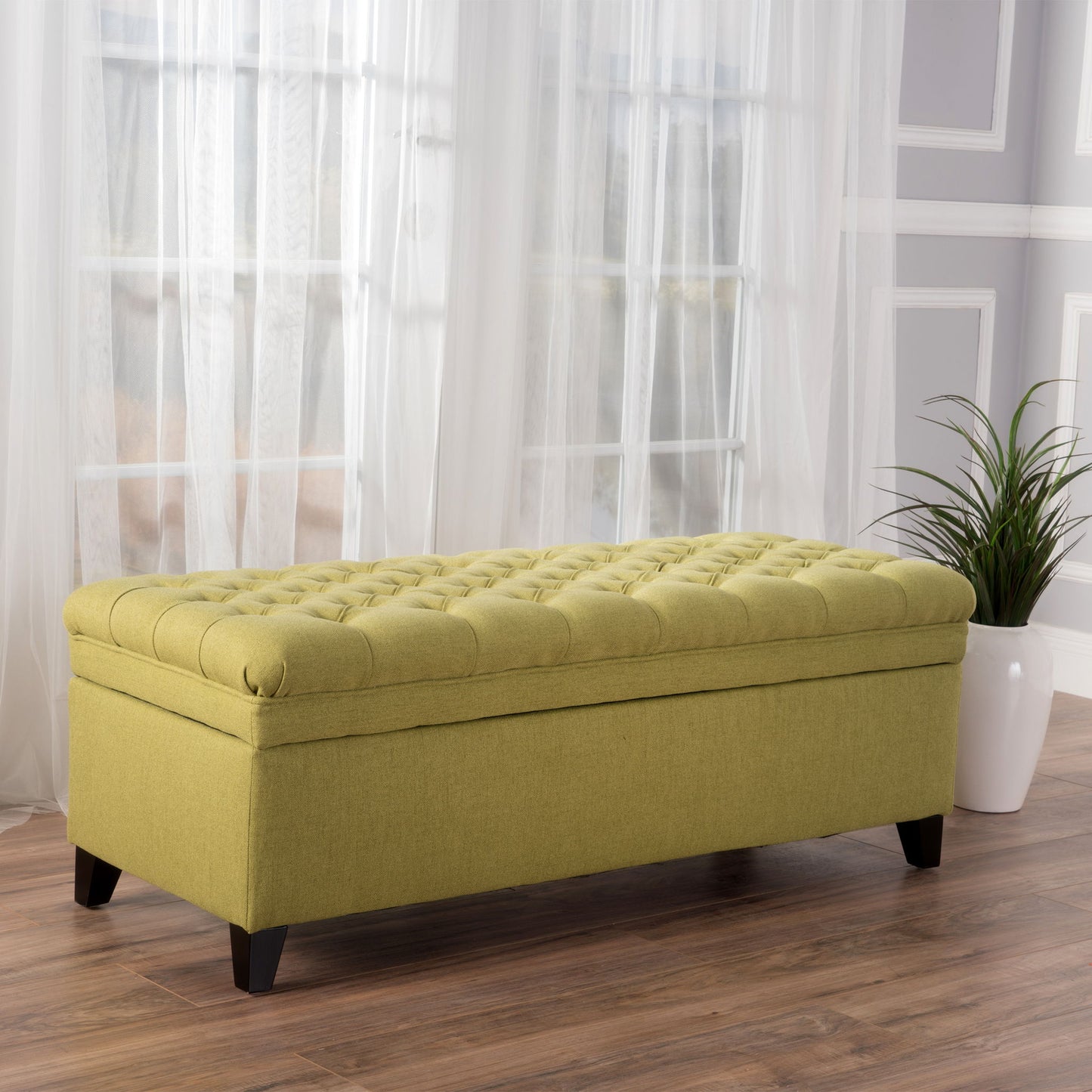 The Juliana Storage Ottoman with birch legs is placed on wooden flooring near tall windows with sheer white curtains and a potted plant in a white vase.