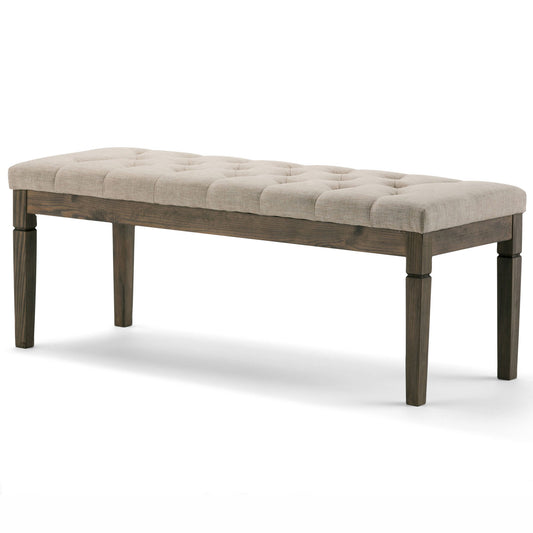 The Waverly Upholstered Tufted Ottoman Bench features a cushioned beige fabric seat with tufting and four dark wooden legs, shown against a plain white background.