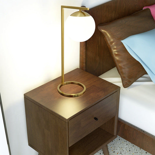 A modern wooden nightstand with a drawer and open shelf holds the Globe Table Lamp Opal With Dimmer Switch Inline in Gold/White, beside a bed with brown pillows and light blue and pink bedding.