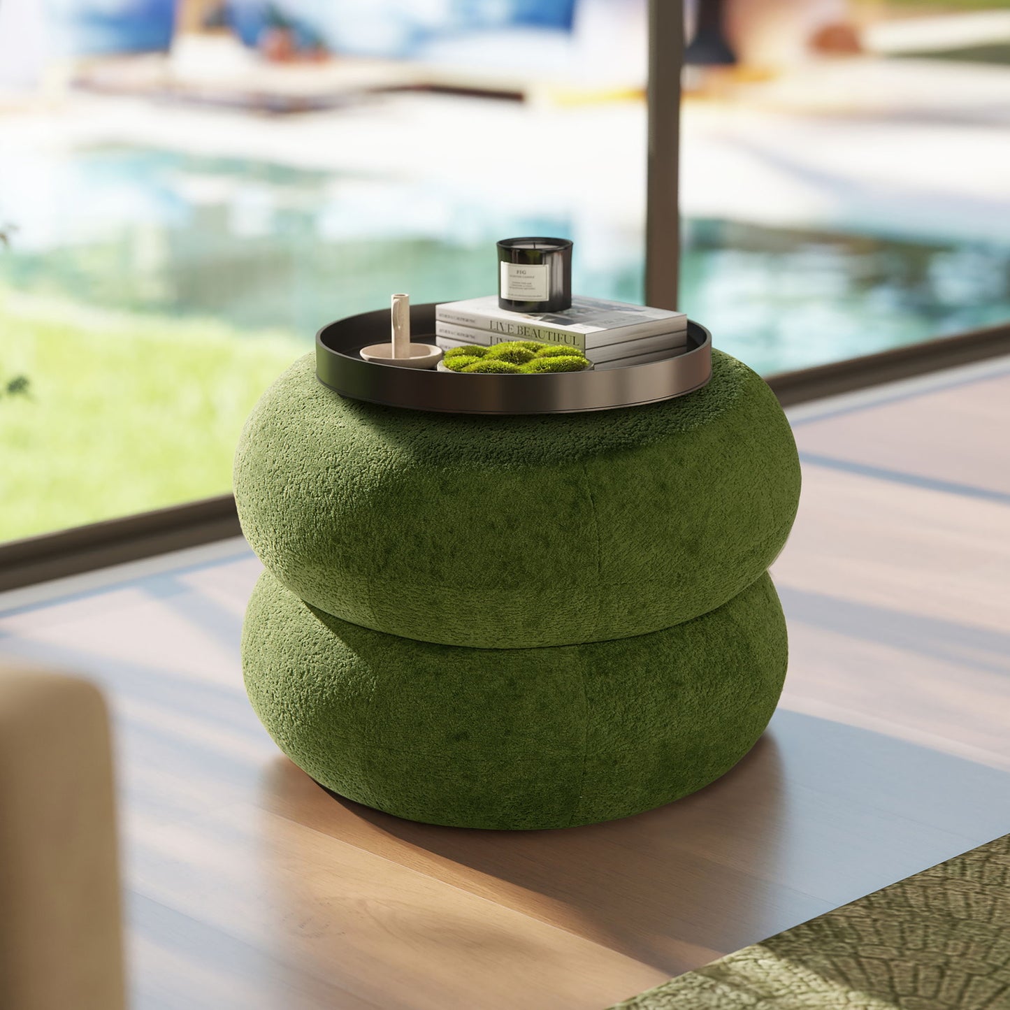 The Nest - Low Round Upholstered Ottoman in green, featuring a textured surface, sits on a wooden floor. A black tray on top holds a candle, books, and decor. Sunlight pours in from large windows beside the pool.