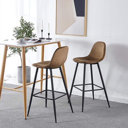 Two Velvet Barstool With Back (Set of 2) in brown with black legs sit by a wooden table adorned with plates, cups, and candles. A plant and abstract art complete the bright, minimalist room.