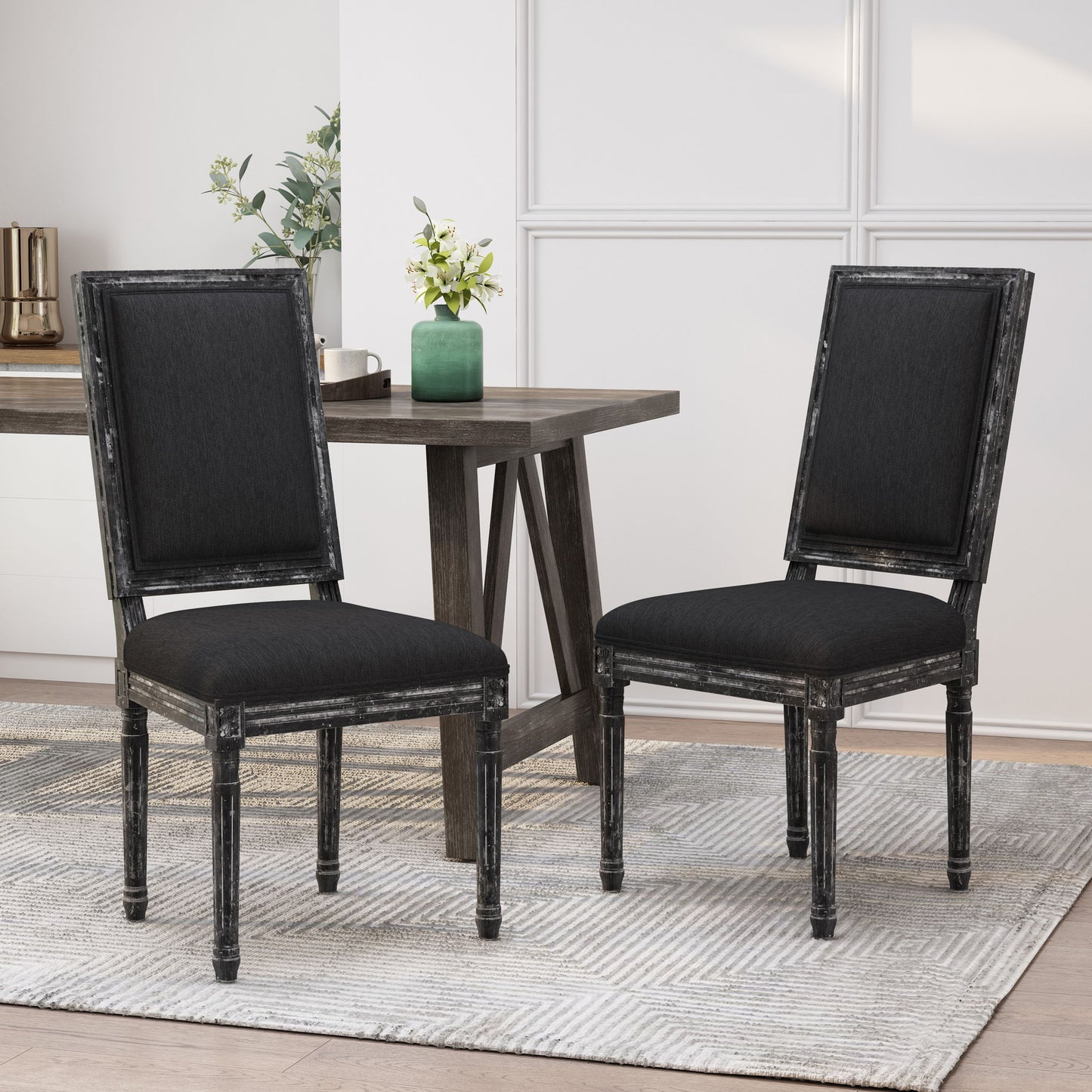 Two French Country Style Dining Chairs with upholstered seats and fluted legs, featuring distressed wooden frames in black, sit on a textured rug beside a dark wood table. A green vase with white flowers decorates the background.