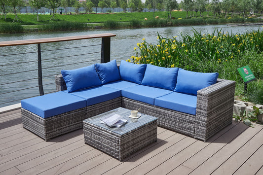 The Patio Sofa With Cushions in gray wicker features blue cushions and comes with a matching coffee table, set on a wooden deck by a river with reeds and yellow flowers nearby. A cup and magazine rest on the table.