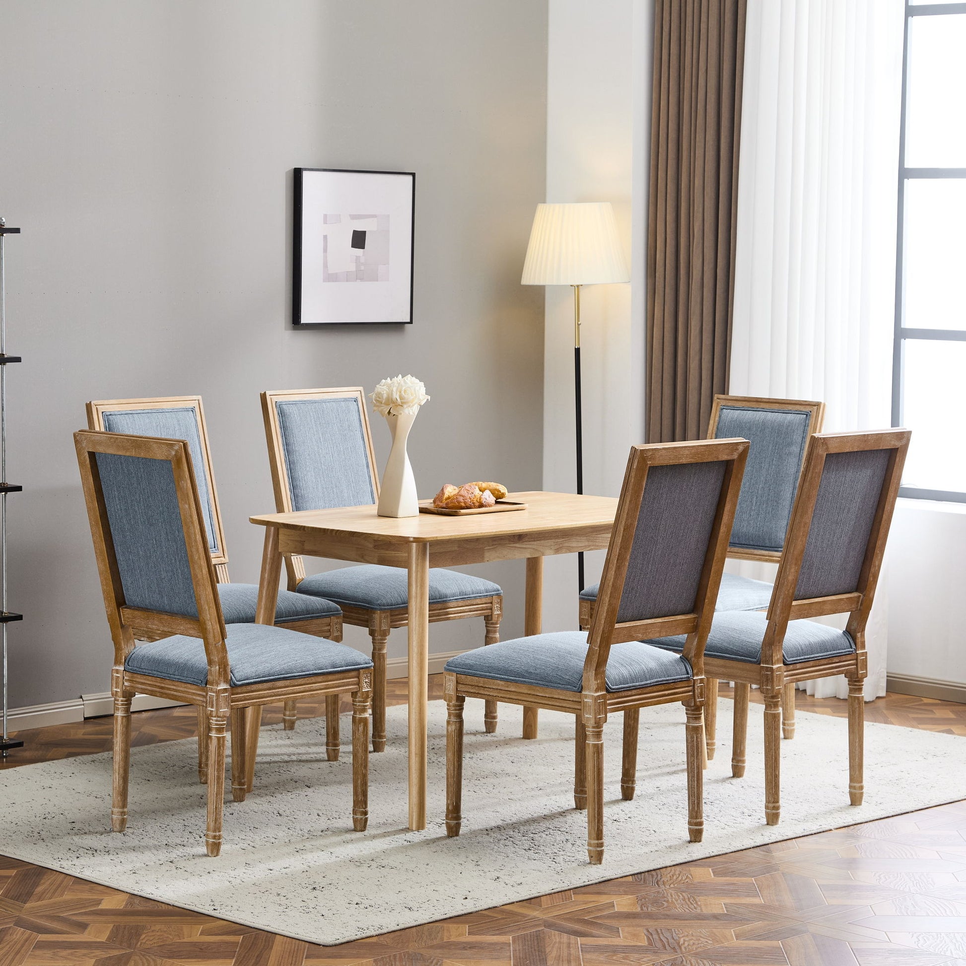 A dining room with a light wood table, six French Country Style Dining Chairs with upholstered seating and fluted legs, a white vase with flowers, pastries on a plate, wall art, a floor lamp, and large curtained windows letting in natural light.