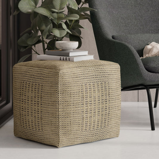 The Delilah Square Woven Pouf in natural serves as a side table, displaying books and a plant beside a modern dark gray armchair with a cushion in a contemporary living area.