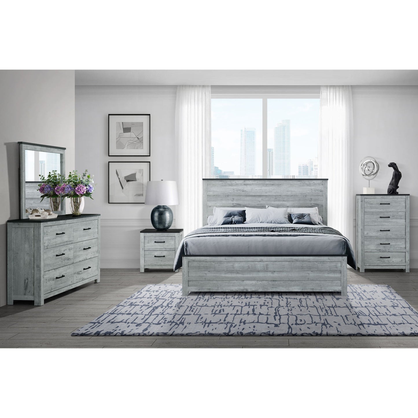 The Kicks - 5 Pieces Bedroom Set features light gray wood furniture: bed, dresser with mirror, nightstand, and tall chest. A rug covers the floor, city views are seen through large windows, and the decor includes flowers and framed art.