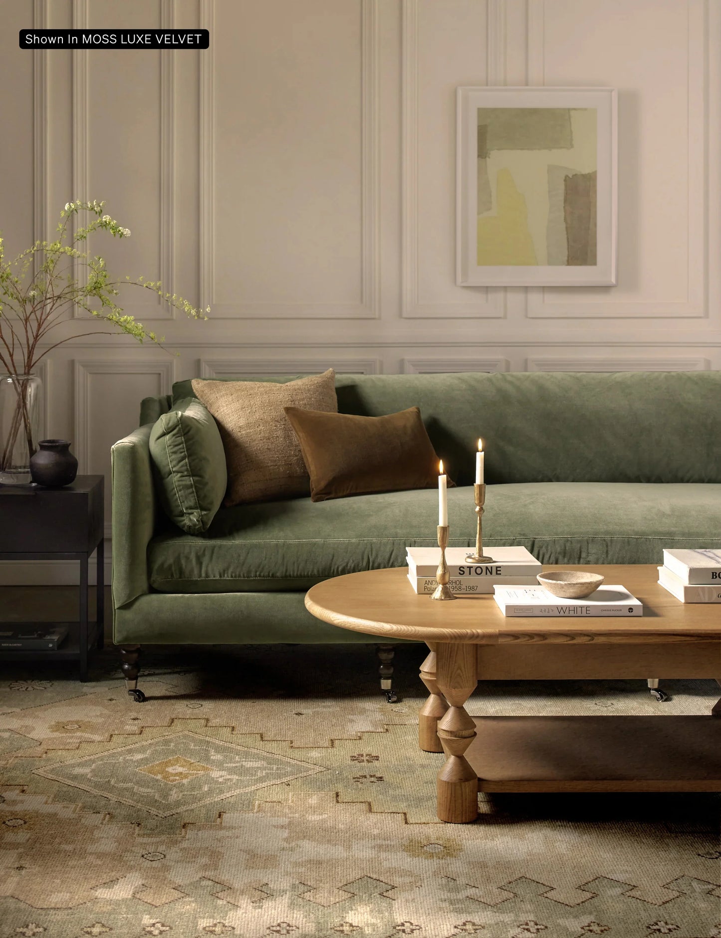 A cozy living room featuring the Fabienne Sofa in green velvet, adorned with brown and beige pillows, complemented by a wooden coffee table. Two lit candles and books rest on the table, while a potted plant and an abstract painting add decorative touches to the space.