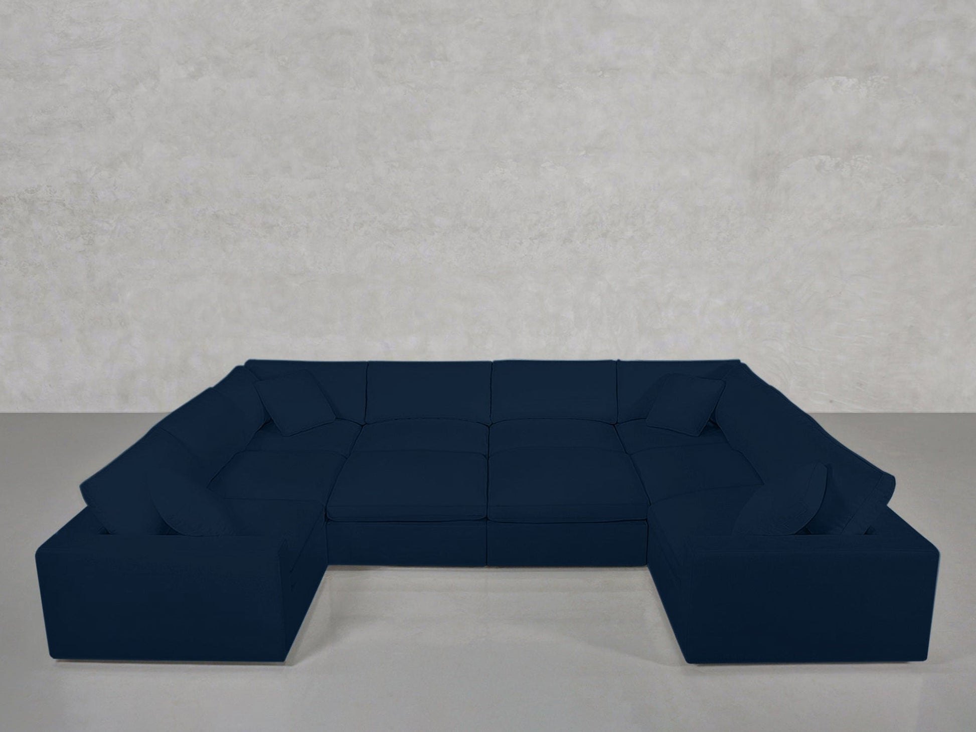 A spacious, dark blue 10-Seat Modular Closed Pit sectional sofa with cushions sits against a plain light gray wall and floor in a minimalistic, empty room.