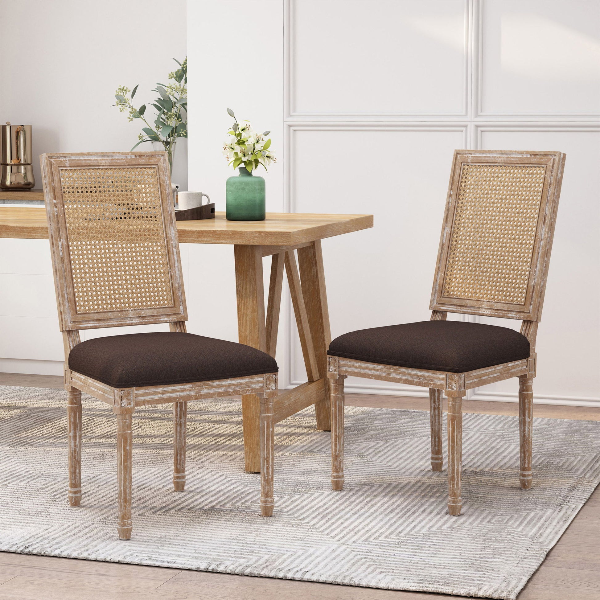 Two French Country Dining Chairs with rectangular cane backrests and smooth upholstered seats are placed by a light wood table. A green vase of flowers sits on the table, and a light rug covers the floor.