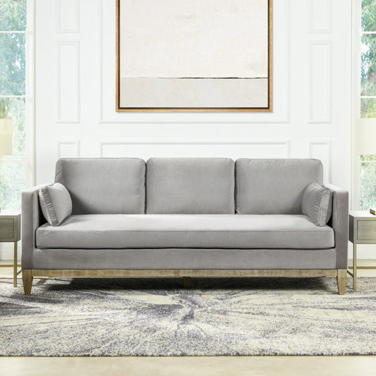 A modern living room features the Knox - Modern Farmhouse Sofa in light gray with two round pillows, set on a patterned rug. White paneled walls, large windows, and a minimalist abstract painting in a gold frame complete the look above the sofa.