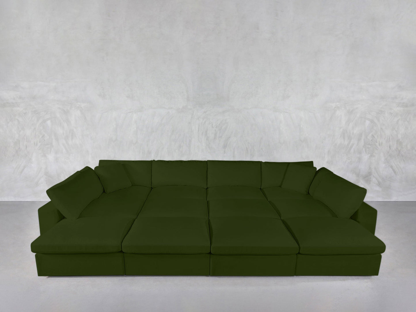 The 12-Seat Deep Modular Daybed, in deep green with multiple seat cushions and back pillows, stands against a light gray textured wall in a minimalistic room with a pale floor.