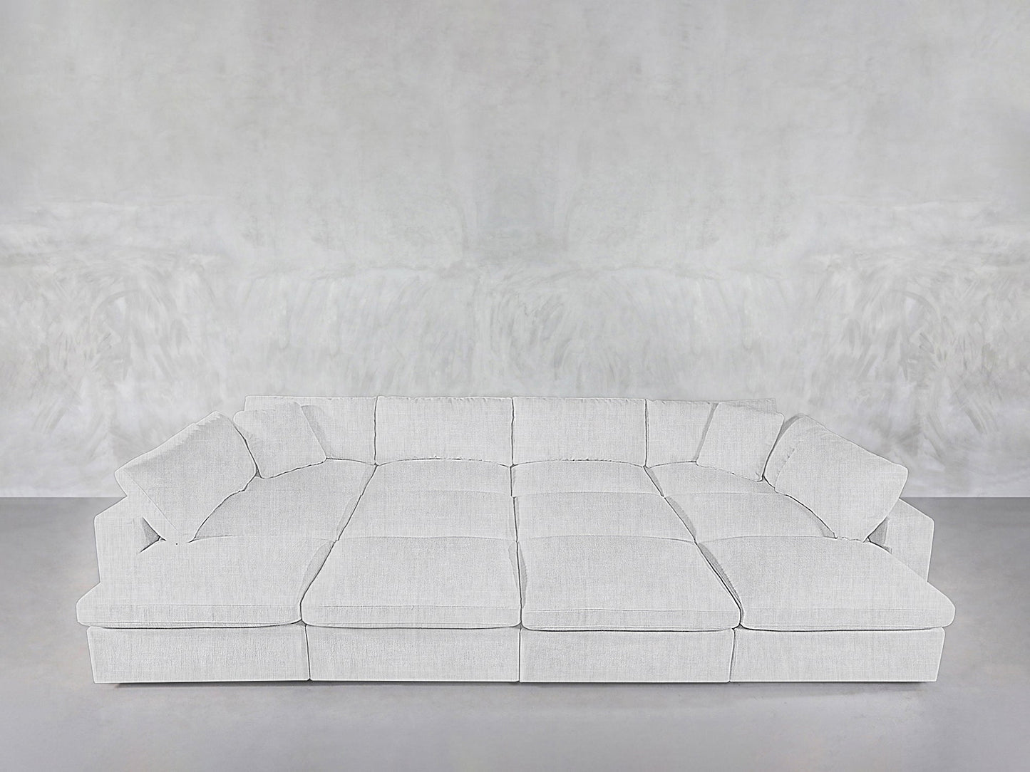 A spacious, modern 12-Seat Deep Modular Daybed with plush cushions and matching pillows is arranged against a plain, light gray textured wall and floor.