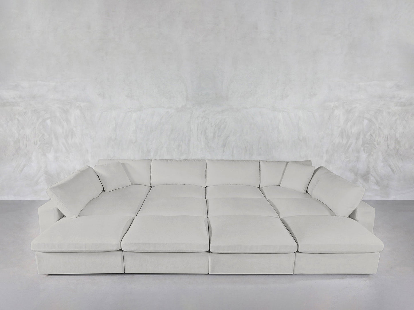 A spacious, modern 12-Seat Deep Modular Daybed with multiple cushions sits against a matching textured gray wall and floor in a minimalist room.