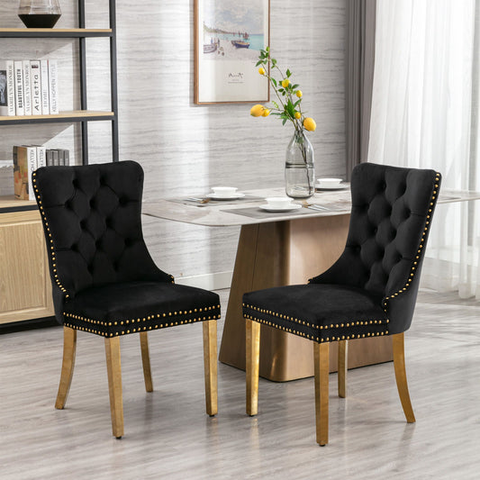 A set of two Nikki black velvet tufted dining chairs with golden stainless steel legs and nailhead trim are featured in a modern dining room with a light wood table, yellow flower vase, sheer curtains, and shelves in the background.