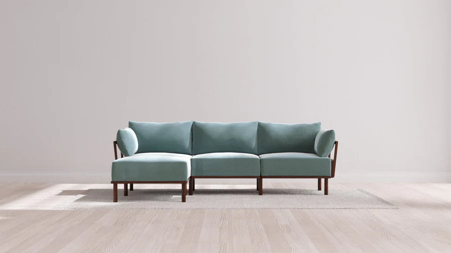 A minimalist, modern three-seater sofa with light teal cushions and wooden armrests sits on a textured rug in a bright room with light wood flooring and plain white walls.