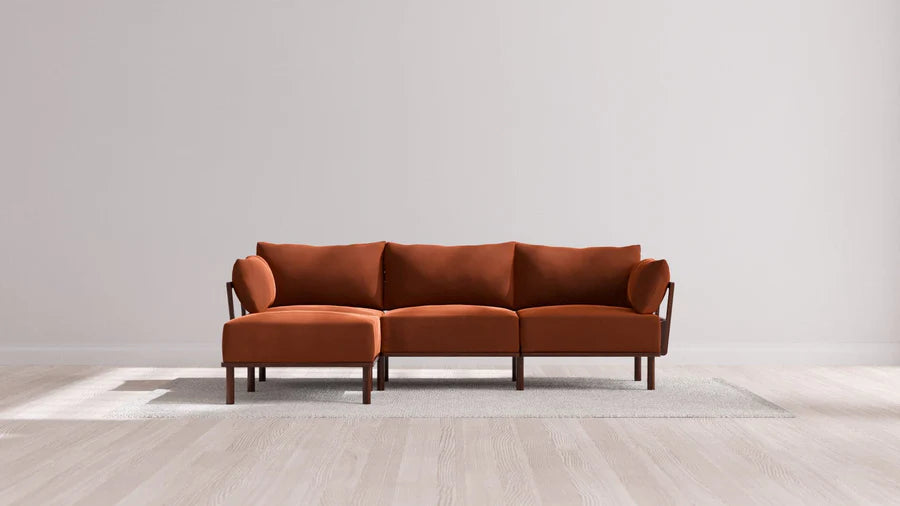 A modern, rust-colored sectional sofa with three seats and a chaise lounge on the left. It's placed on a light-colored rug against a plain, off-white wall in a spacious, well-lit room with light wooden flooring.