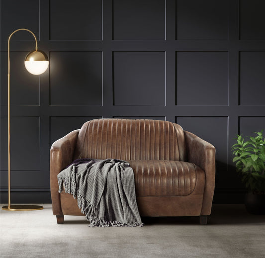 A modern living room showcases the Brancaster Grain Leather Loveseat in brown with a gray knitted throw, a gold floor lamp with a round white shade, dark paneled wall, and a potted plant on the right.