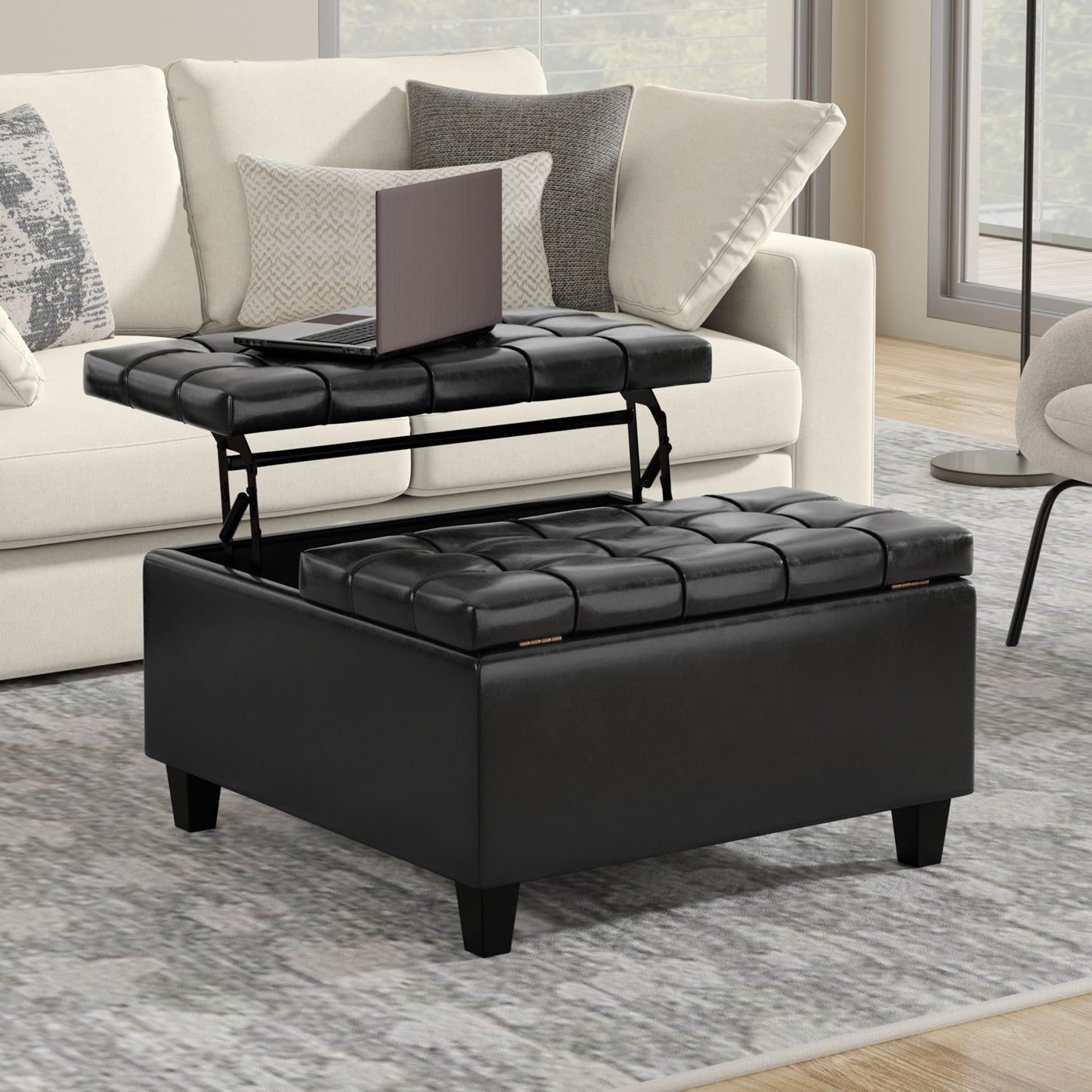 The Harrison Upholstered Coffee Table Storage Ottoman with a black tufted lift-top tray holding a laptop is placed on a gray rug in front of a beige sofa with patterned pillows in a modern living room.