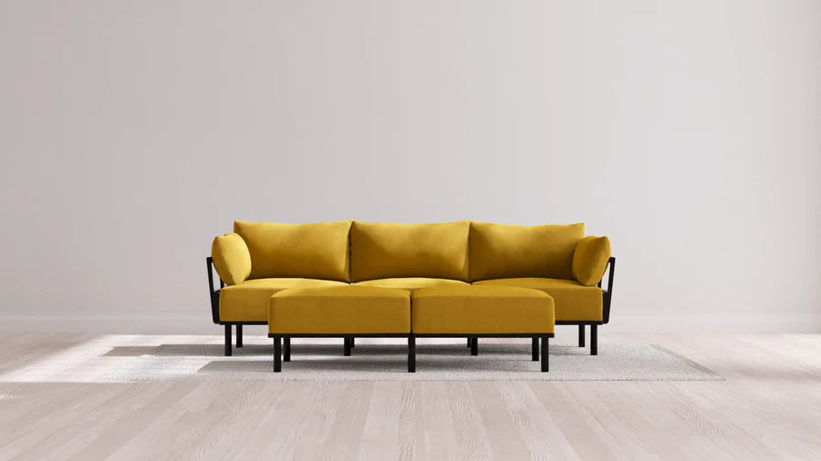 A modern balsa-colored Jack 3-Seater Sofa with a matching Bench Ottoman is placed on a light gray rug. The sofa features black metal legs and is set against a plain white wall, while wooden flooring can be seen in the foreground.
