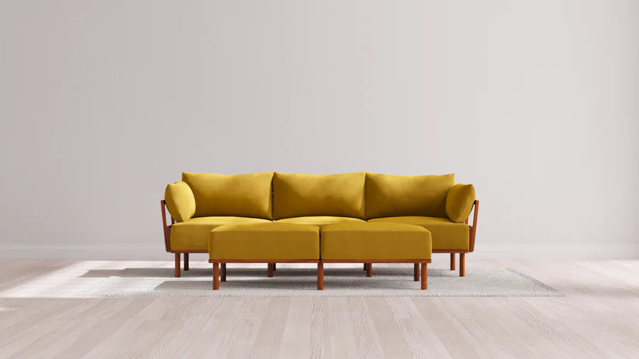 The Indoor Jack 3-Seater Sofa + Bench Ottoman in Balsa, with its mustard yellow hue and wooden legs, is placed atop a light gray rug in a minimalist room characterized by a light wood floor and plain off-white walls. The sofa showcases clean lines and comes with two matching bolster pillows.