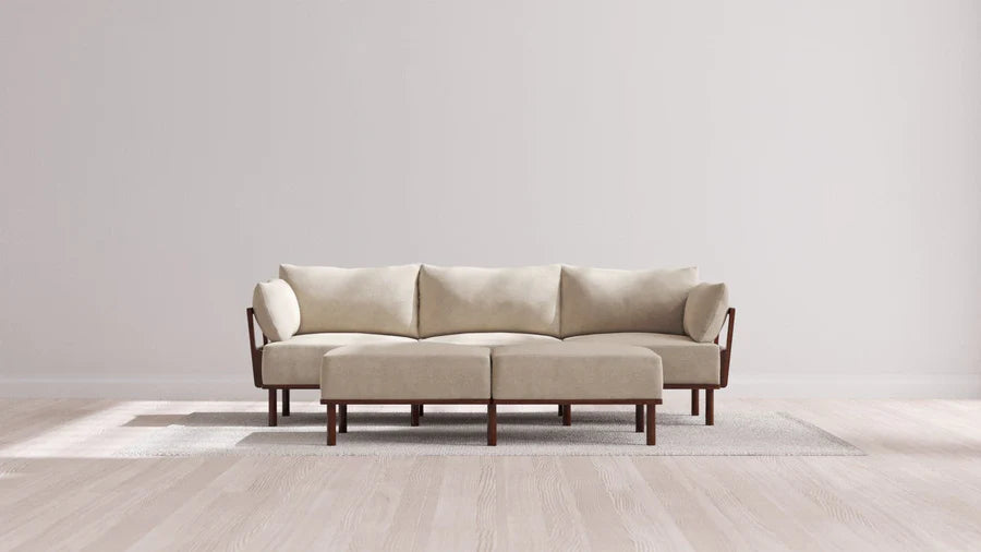 An Indoor Jack 3-Seater Sofa with a Bench Ottoman in Balsa is placed on a light-colored rug in a bright room featuring wooden flooring and plain walls. The sofa's wooden arms and legs enhance the neutral, modern aesthetic.