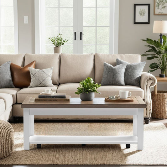 A cozy living room with a beige sofa, gray and brown pillows, and the Hampton Coffee Table in Jasmine Whitewash/Barnwood, topped with books, a plant, and mugs on a woven rug by large windows streaming natural light.