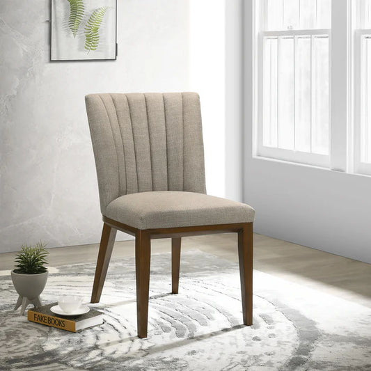 An Elmwood Stylish Dining Chair (Set of 2) with beige upholstery and wooden legs sits on a round patterned rug near a sunlit window. Nearby are two books labeled "FAKE BOOKS," a small plant, and a framed fern print on the wall.