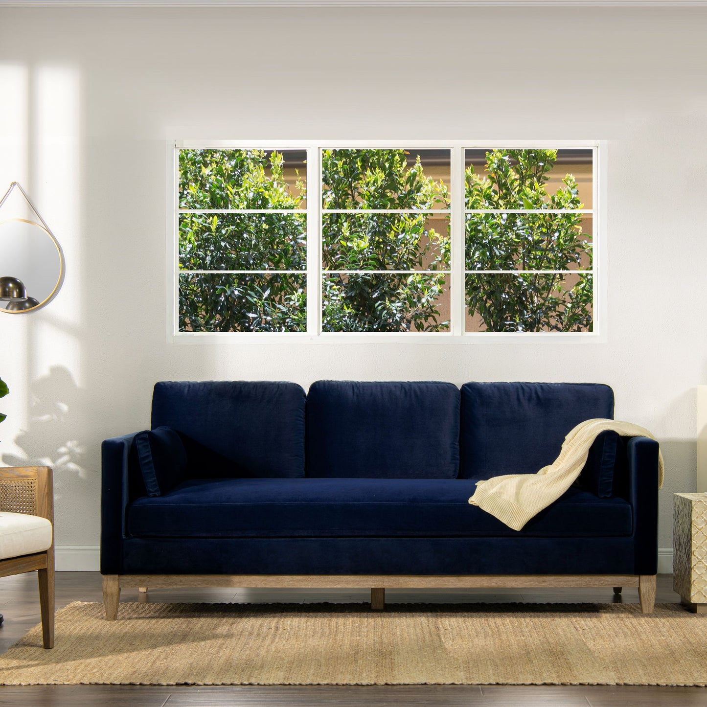 A modern living room features the Knox - Modern Farmhouse Sofa in dark blue velvet, a cream throw blanket, a beige chair, a round wall mirror, a jute rug, and large windows overlooking lush green trees.