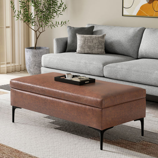 The Rebecca Multifunctional Coffee Table Storage Ottoman With Lift Up Lid, featuring brown leather and black legs, holds a tray with a cup and glasses before a modern light gray sofa with patterned pillows in a bright living room.