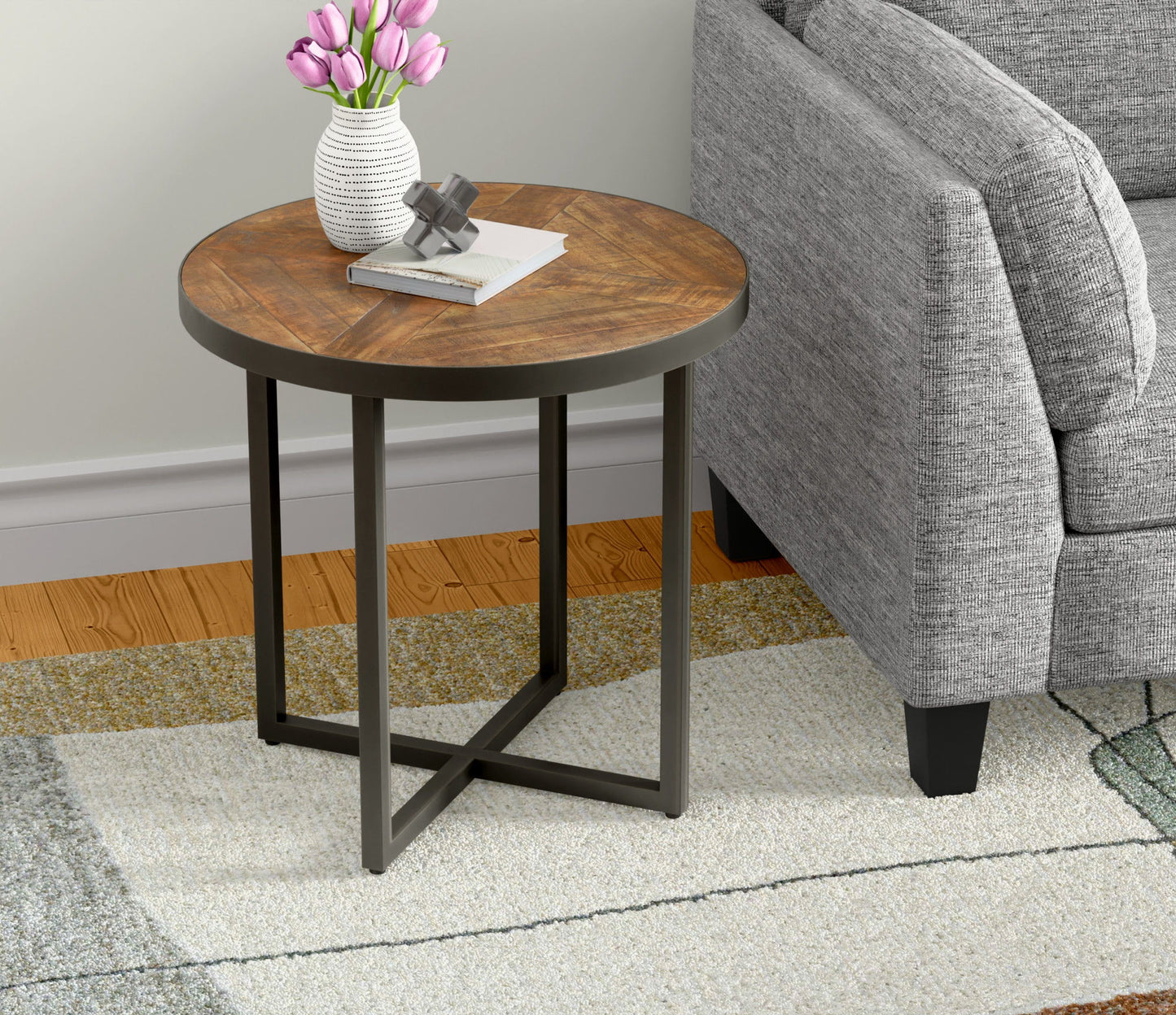 The Faro Round Table with metal legs stands by a gray sofa. On it are a white vase of pink tulips, a closed book, and a small silver ornament, set atop a patterned rug.