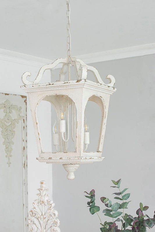A 4 Light Wood Chandelier in cream white with an adjustable chain is suspended from the ceiling. The hanging light fixture, bulbs not included, features a vintage look with greenery and ornate decor softly visible in the background.