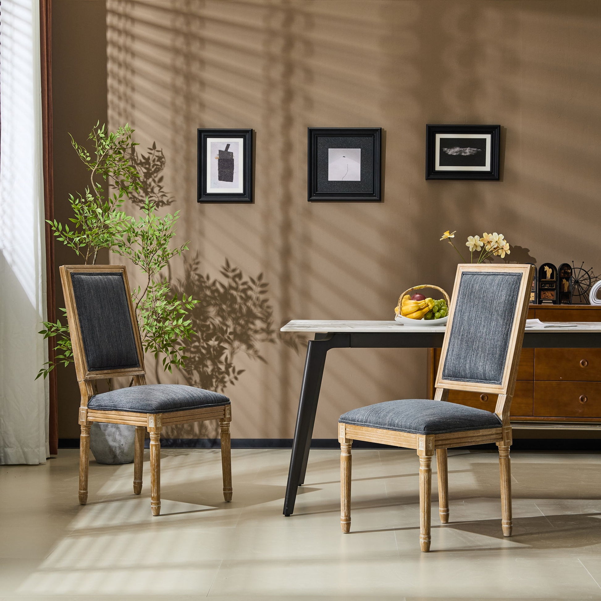 Two French Country Style Dining Chairs with upholstered seats and fluted legs sit near a small table with a fruit bowl. A green plant, framed wall art, and a decorated sideboard complete the cozy, sunlit room.