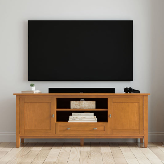 A flat-screen TV is mounted on a white wall above the Warm Shaker - TV Media Stand - Brown, which features drawers, cabinets, books, a small plant, a decorative box, and a microphone on top. The floor is light wood.
