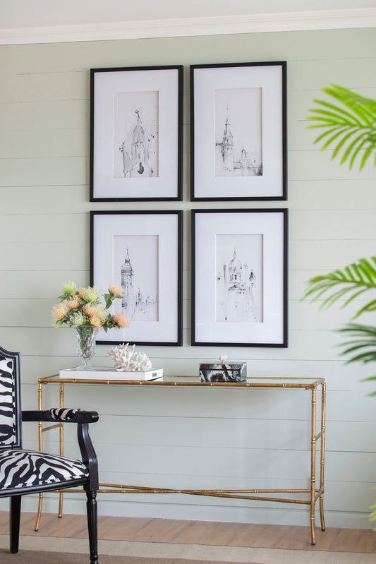 A modern living space features the Architecture Wall Art Prints (Set of 4, White/Black) on a light green shiplap wall, paired with a glass console table with gold legs, floral decor, and a zebra-patterned chair.