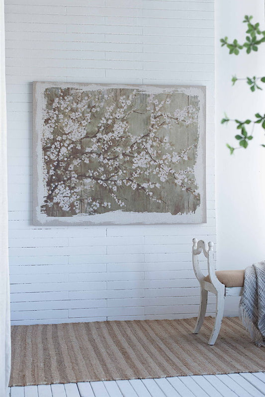 A minimalist room with white paneled walls features a Large Cherry Blossom Canvas Art Print in gray and white matte, a vintage chair with a tan seat draped with a gray blanket, a beige striped rug, and green leaves in the foreground.