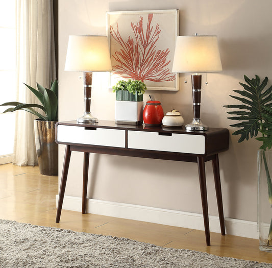 The Christa Sofa Table in White/Espresso features two drawers and displays matching lamps, a red teapot, a plant, and a decorative bowl. Above it hangs coral artwork, flanked by green plants on both sides with a beige rug below.