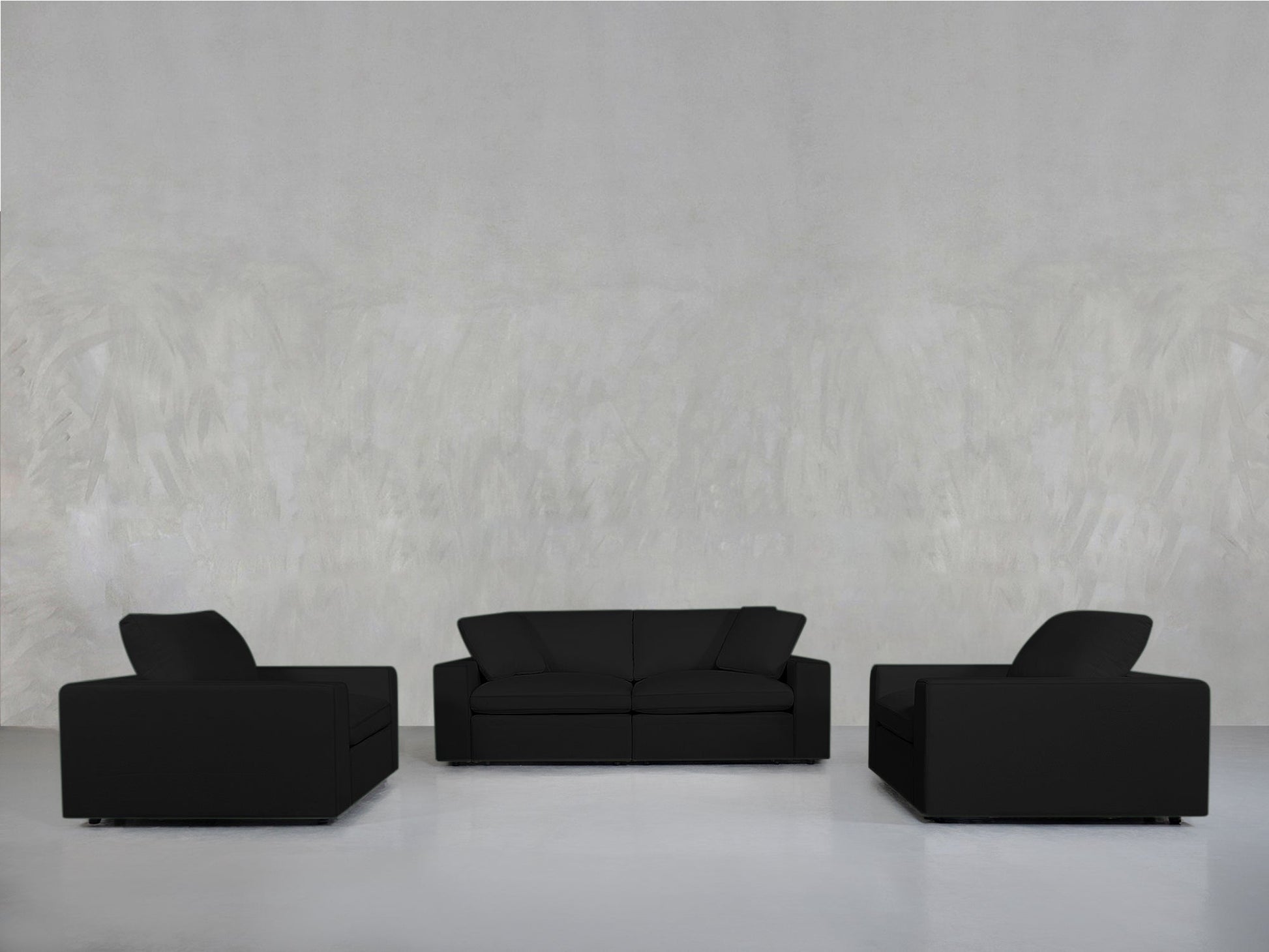 A minimalist room with a light gray wall and floor showcases the 2-1-1 Sofa & Armchair Set: a black sofa centered between two matching black armchairs, all symmetrically arranged and facing inward.