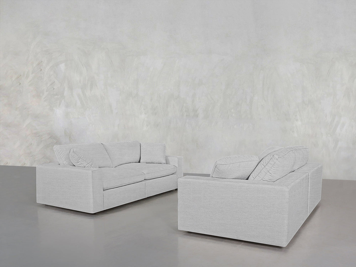 The 2-2 Sofa Set, featuring two light gray sofas with loose back cushions, is arranged facing each other in a minimalist room with a gray floor and a textured, light-colored wall.