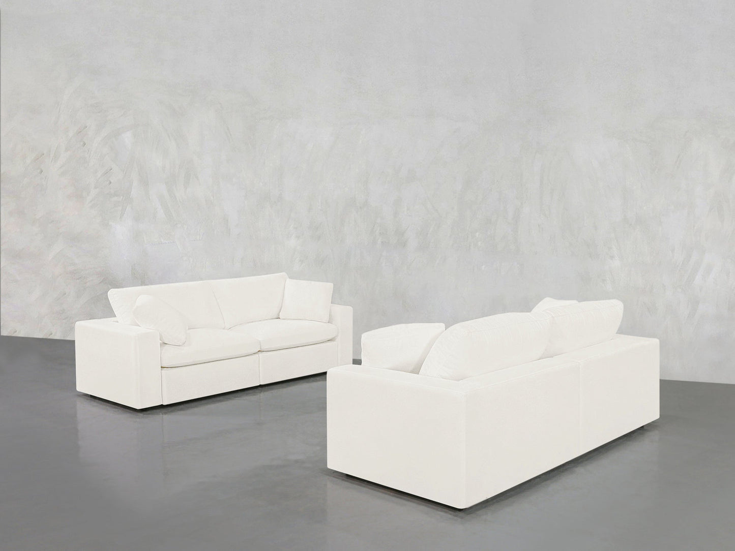 The 2-2 Sofa Set, with its modern white design and plush cushions, is arranged apart on a shiny gray floor against a textured light gray wall in a spacious, minimalist room.