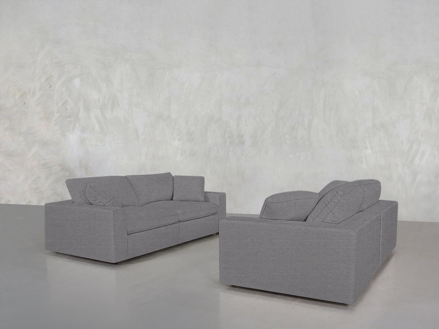 The 2-2 Sofa Set, featuring modern gray sofas with large cushions, is arranged on a smooth, light gray floor against a textured gray wall for a minimalistic, spacious look.