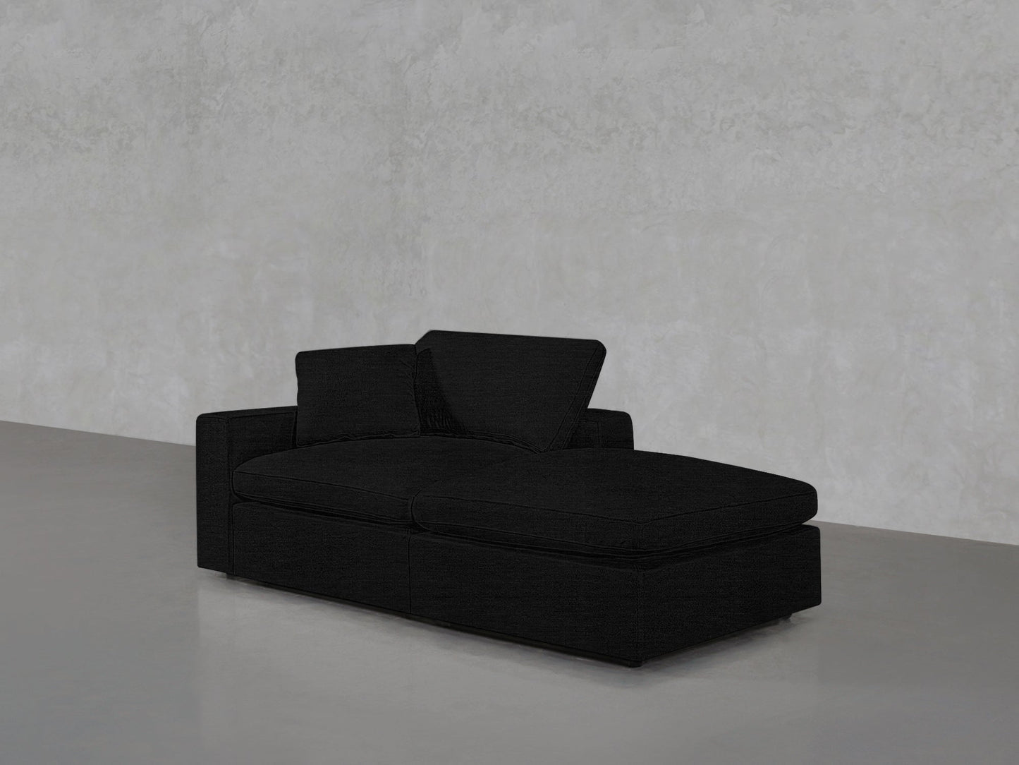 A 2-Seat Modular Lounger Loveseat in black, featuring two cushions and a chaise lounge, is set against a light gray textured wall on a smooth gray floor.