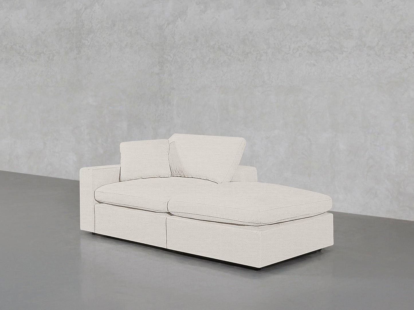 A 2-Seat Modular Lounger Loveseat in light beige with two matching cushions is set against a textured light gray wall and polished gray floor.