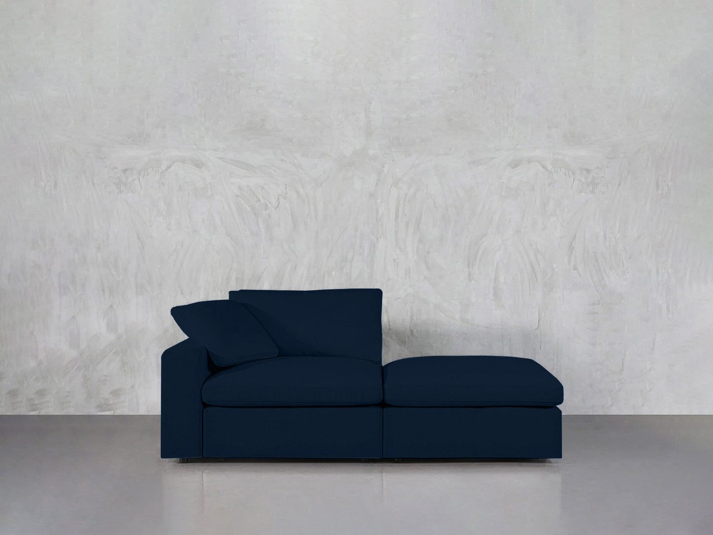 A 2-Seat Modular Lounger Loveseat in dark blue with a left armrest and matching cushion is placed against a light gray textured wall on a smooth gray floor.