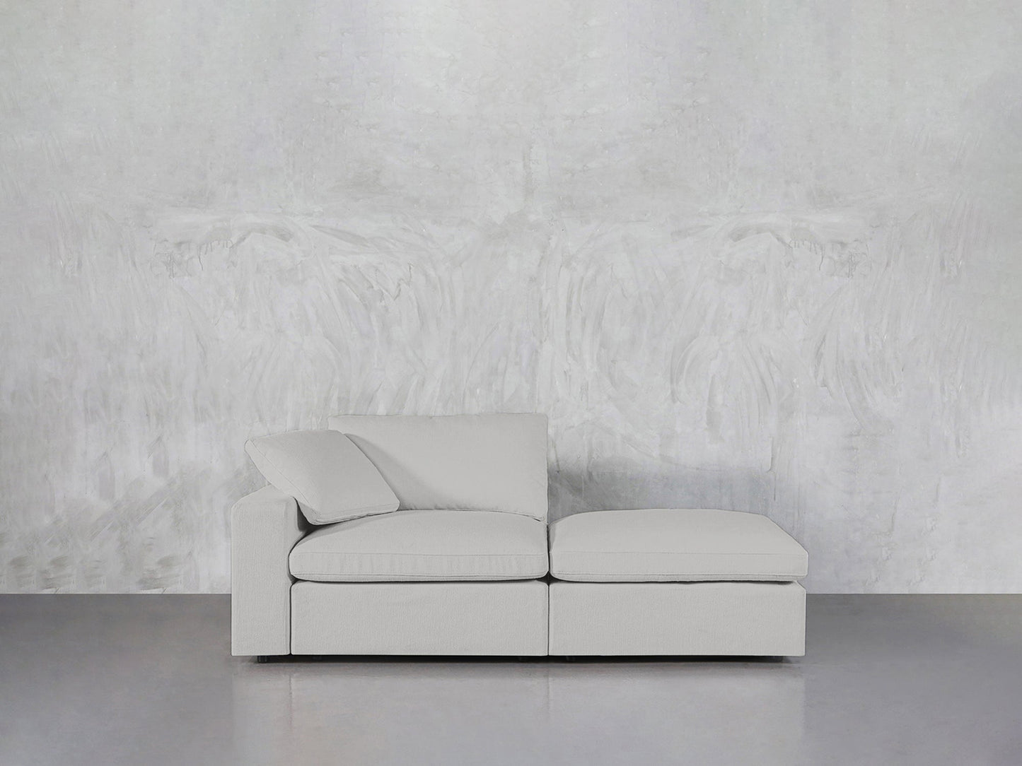 The 2-Seat Modular Lounger Loveseat features clean lines and plush cushions in a modern, light gray design with a left armrest. It sits against a textured, pale gray wall on a smooth, gray floor for a minimalist look.