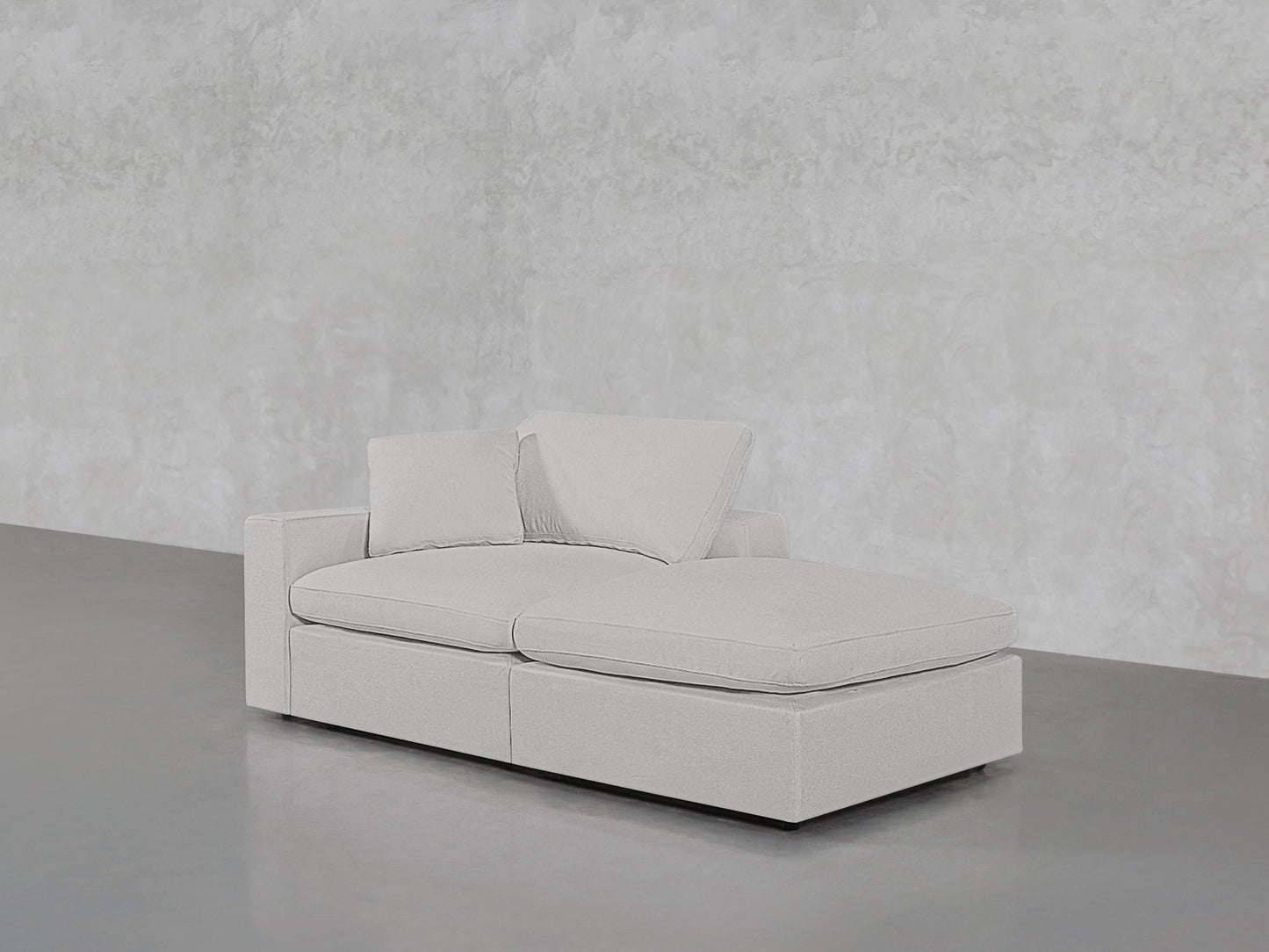 A 2-Seat Modular Lounger Loveseat in light gray, featuring rectangular cushions and two pillows, is set against a textured, light gray wall on a smooth gray floor.