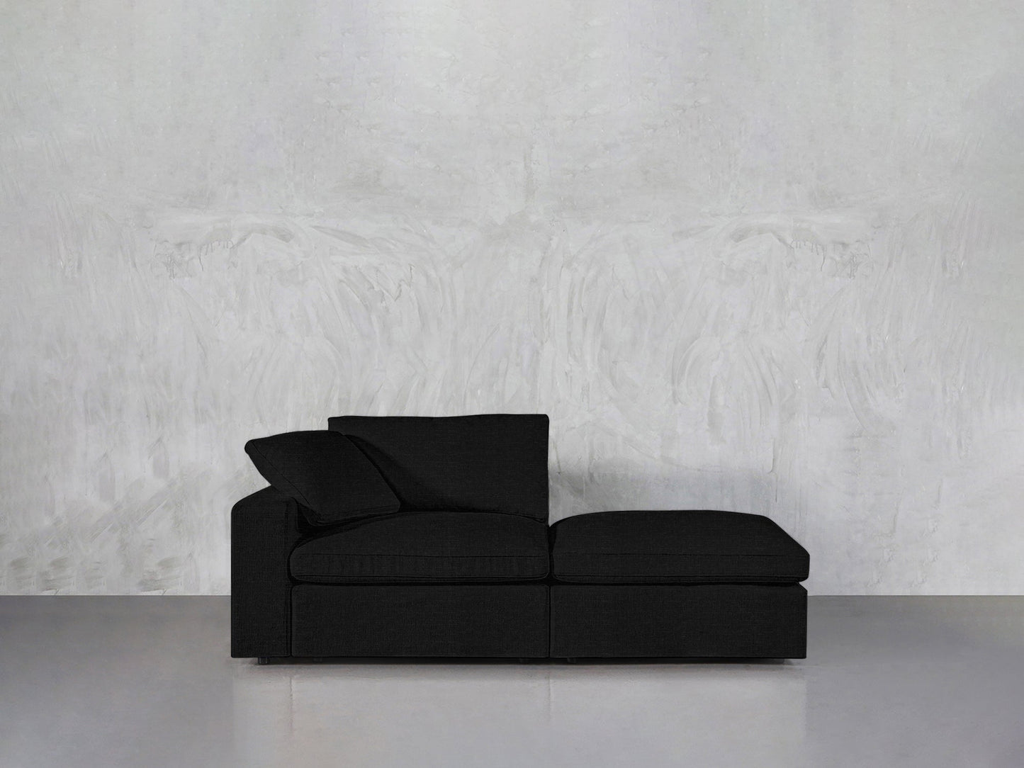 The 2-Seat Modular Lounger Loveseat, featuring a modern black design with a left armrest and two cushions, is set against a light gray textured wall on a smooth gray floor.