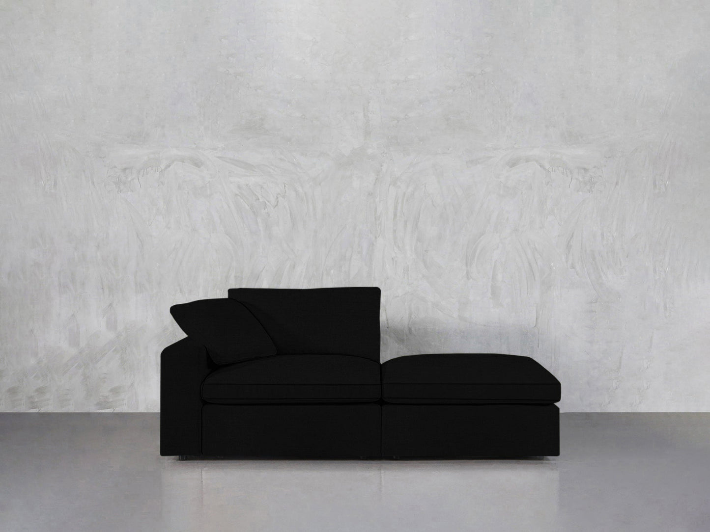 A 2-Seat Modular Lounger Loveseat in black with a pillow is placed against a textured light gray wall on a smooth gray floor.