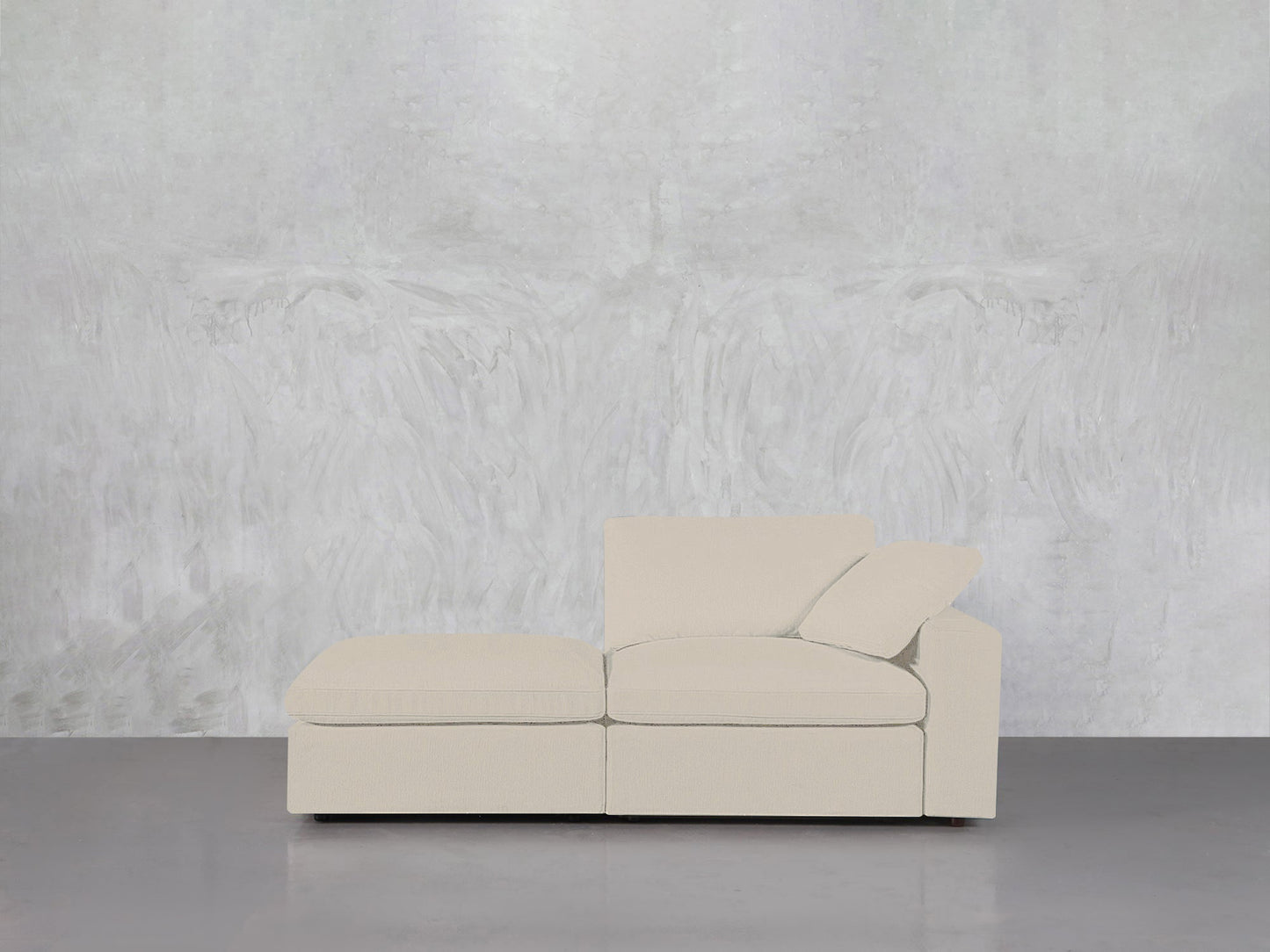 A contemporary 2-Seat Modular Lounger Loveseat in light beige with a chaise lounge sits on a gray floor against a textured light gray wall, featuring one matching cushion on the right side.