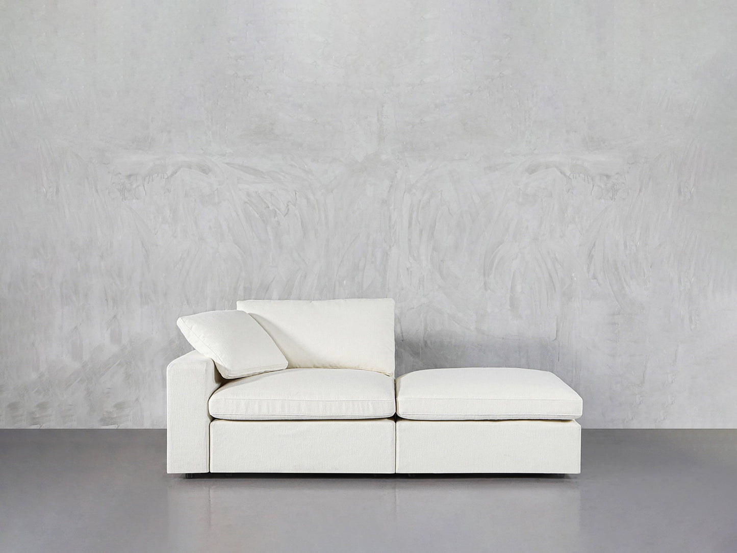 The 2-Seat Modular Lounger Loveseat, featuring clean lines and plush off-white cushions, sits against a textured light grey wall on a smooth grey floor.