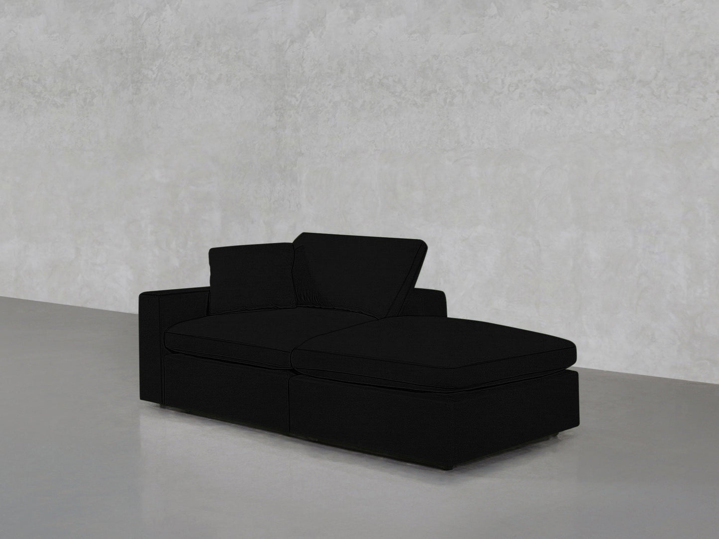 A 2-Seat Modular Lounger Loveseat in black with a right-side chaise rests in a minimalist room featuring a light gray textured wall and gray flooring.